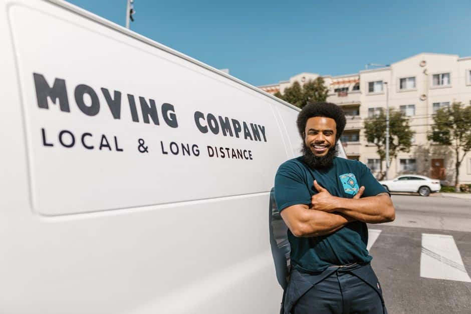 pexels-photo-7464643 - EZ Movers | New Orleans Moving Company Smiling man stands beside a white moving van that reads 'MOVING COMPANY LOCAL & LONG DISTANCE'.