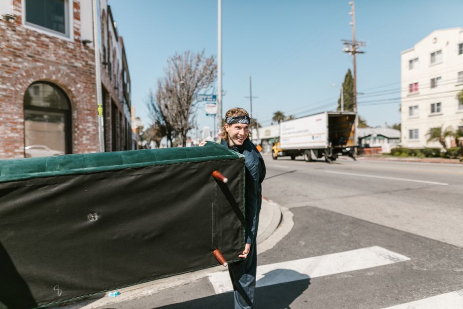 pexels-photo-7464712 - EZ Movers | New Orleans Moving Company Person in a bandana pushes a large black dumpster along a sunny city street by a brick building near a crosswalk.